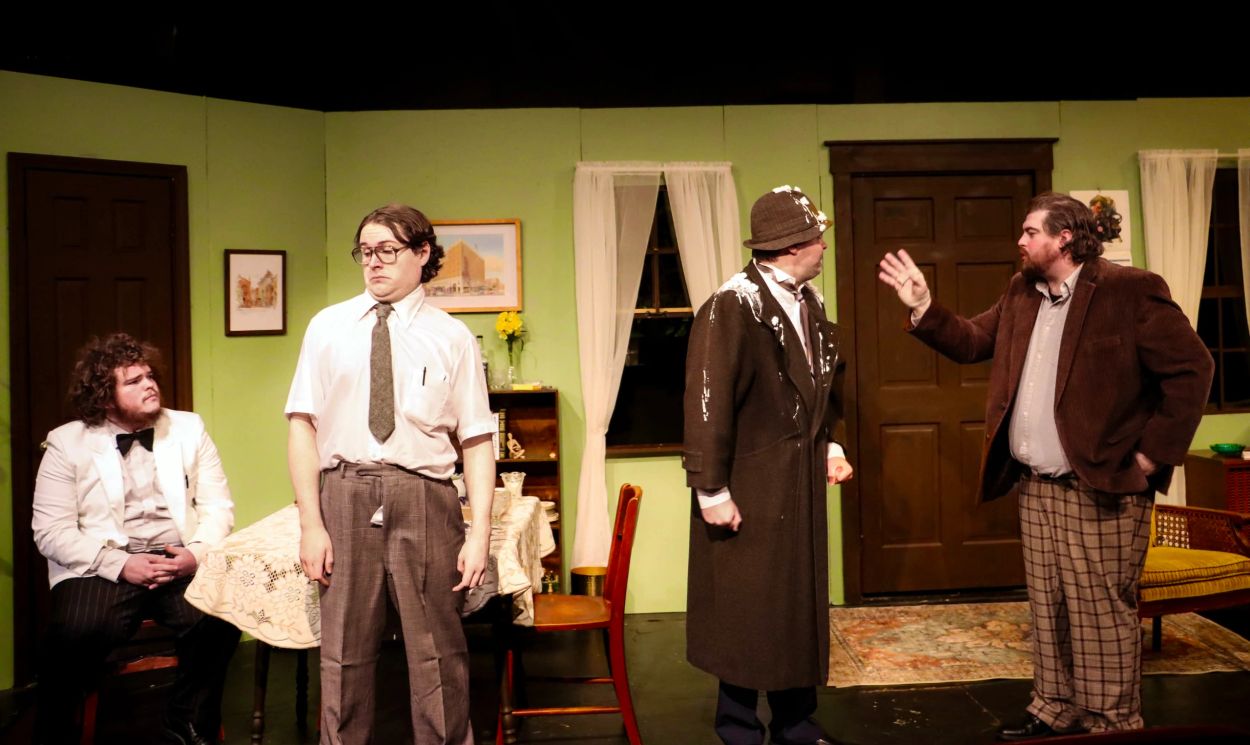 Four men perform a dramatic scene on a stage with vintage decor.