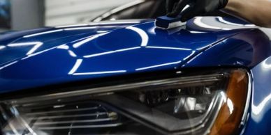 Close-up of a blue car's headlight with a hand cleaning it.