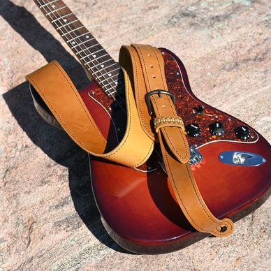 Sunlit electric guitar with a brown leather strap on stone surface.
