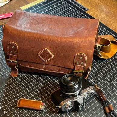 Vintage leather camera bag with Nikon camera and leather-laced lighter on a cutting mat.
