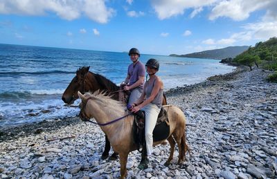 Two people horseback riding on a rocky beach.