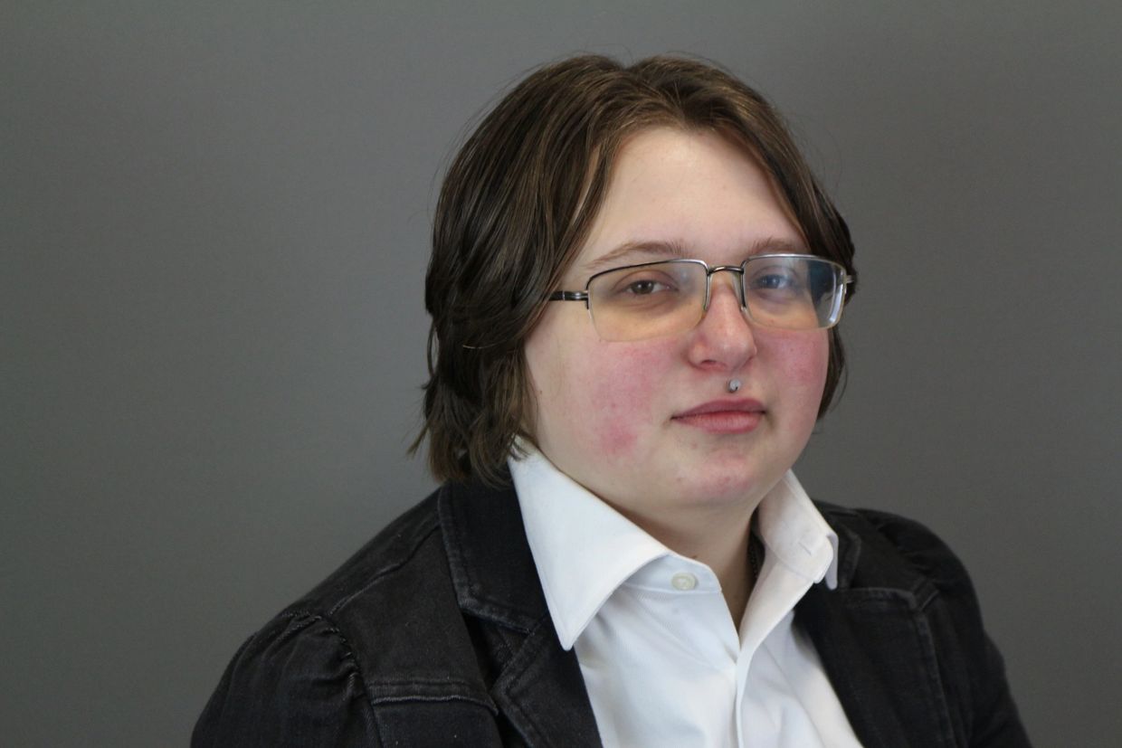 Person with glasses, white shirt, and black jacket against a gray background.