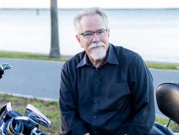Older man in glasses and black shirt near a motorcycle.