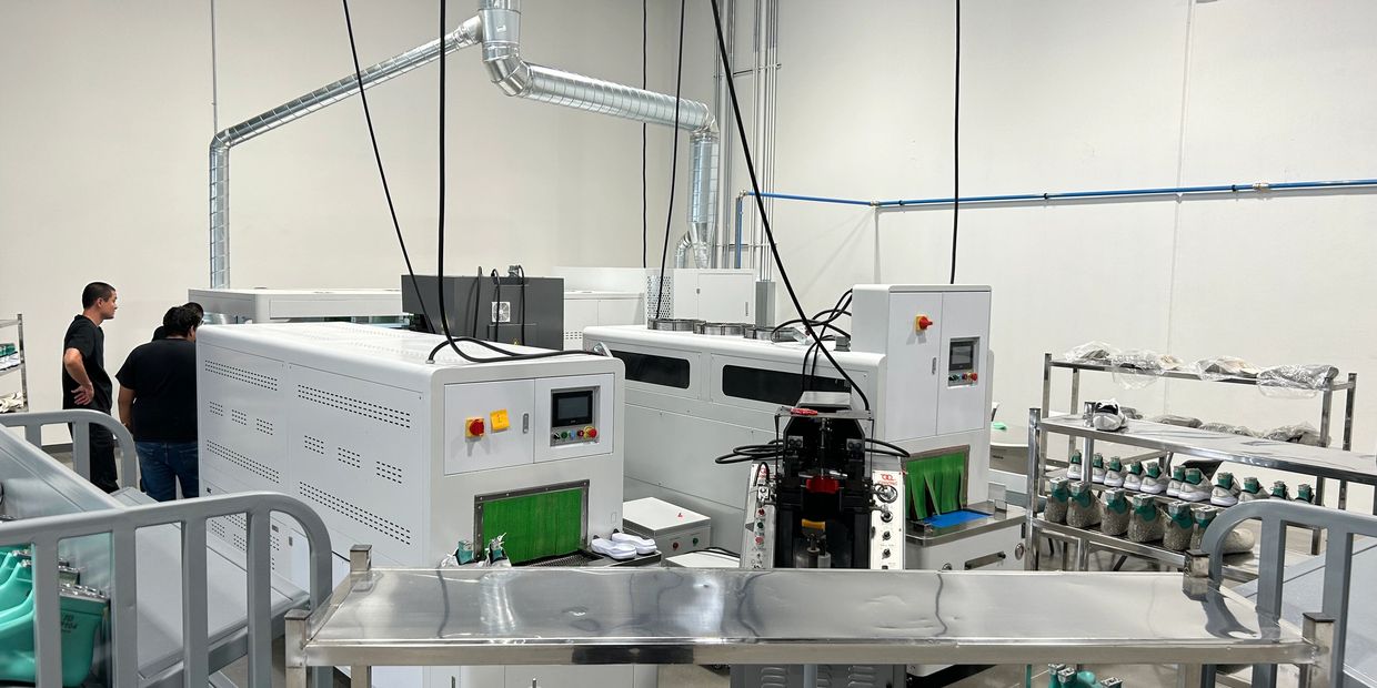 Industrial shoe manufacturing machines with green shoe molds and two people observing.