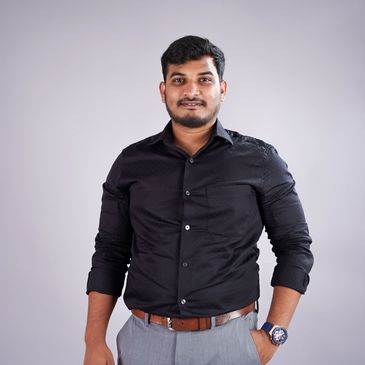 Confident man in black shirt and gray pants posing against a plain background.