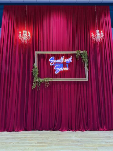 Elegant red curtain backdrop with chandeliers and neon sign framed by greenery.