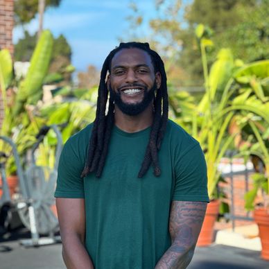 Smiling man with dreadlocks stands outdoors with plants in background.