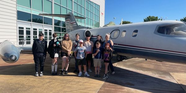 A group of kids and adults posing by a small private jet outside a building.