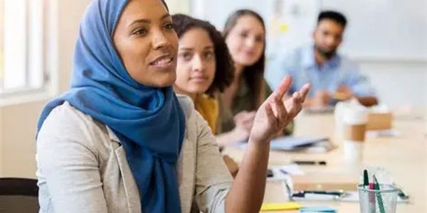 A woman in a blue hijab speaking during a meeting with colleagues.