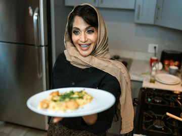 A woman in a headscarf happily presents a plate of food in a kitchen.