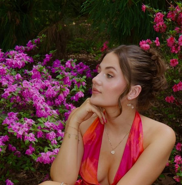 Woman in a vibrant red dress surrounded by pink flowers, gazing thoughtfully.
