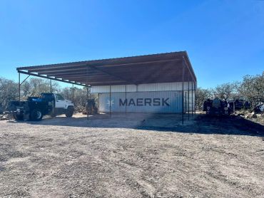 Maersk shipping container under a metal roof with trucks nearby.