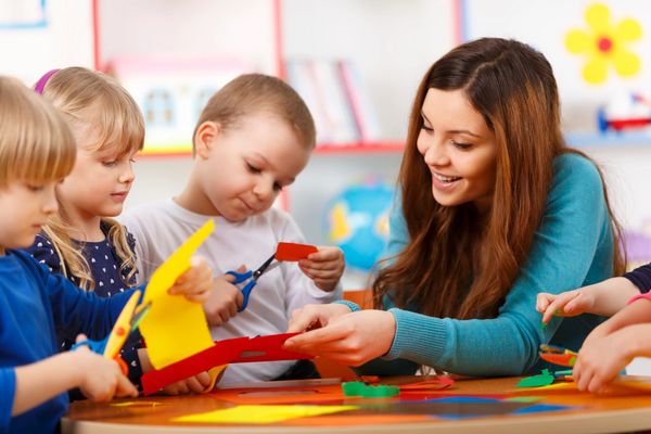 A smiling teacher helps three children with a craft.