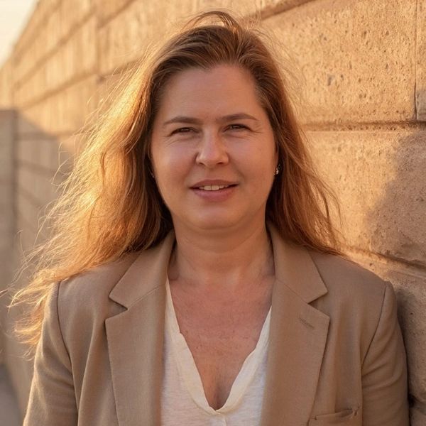 The Valley PMM owner, Lana Bazan, with long hair wearing a beige blazer smiles near a brick wall in sunlight.