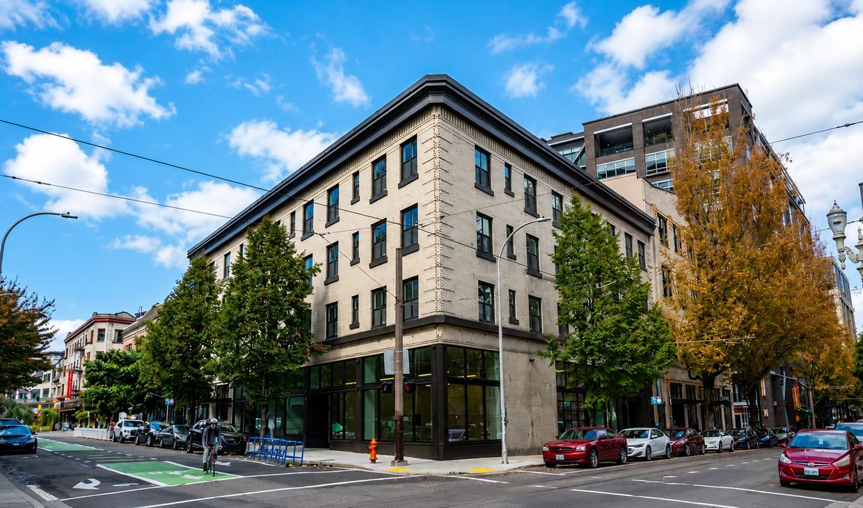 Urban street corner with cars, trees, and a three-story building under a blue sky.