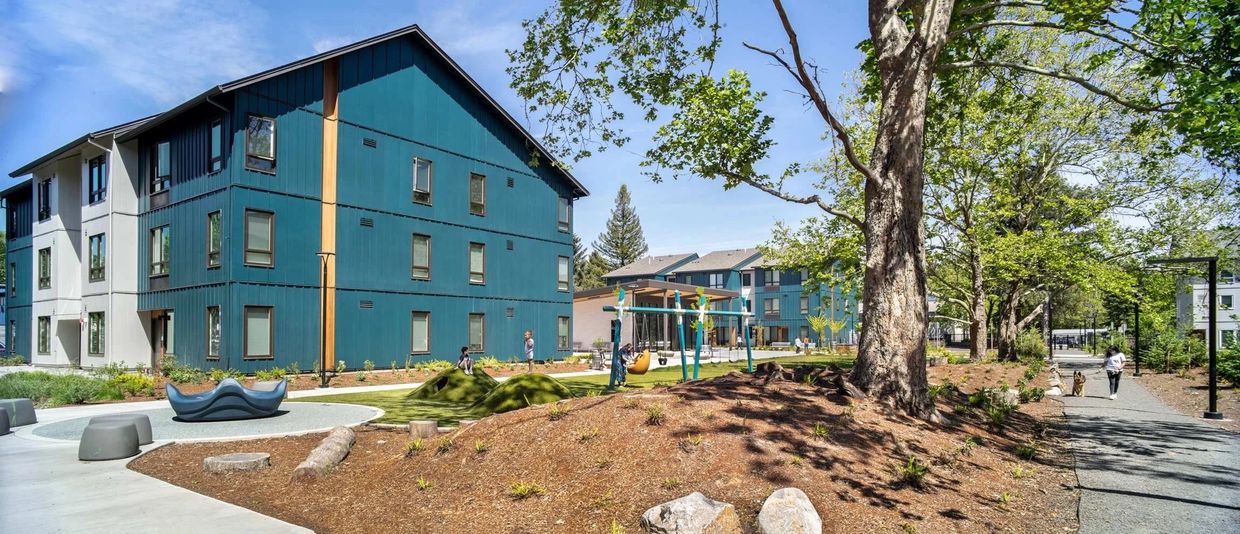 Modern blue apartment buildings with a playground and walking path surrounded by trees.
