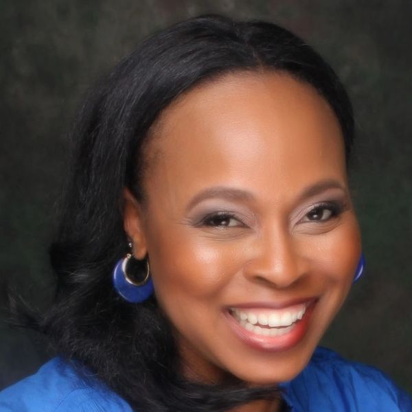 Smiling woman with blue earrings and blouse against a dark background.