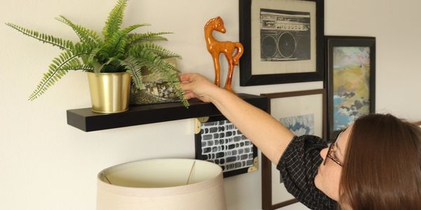 Laura places an accessory on a floating shelf to complete a vignette with framed art and a plant