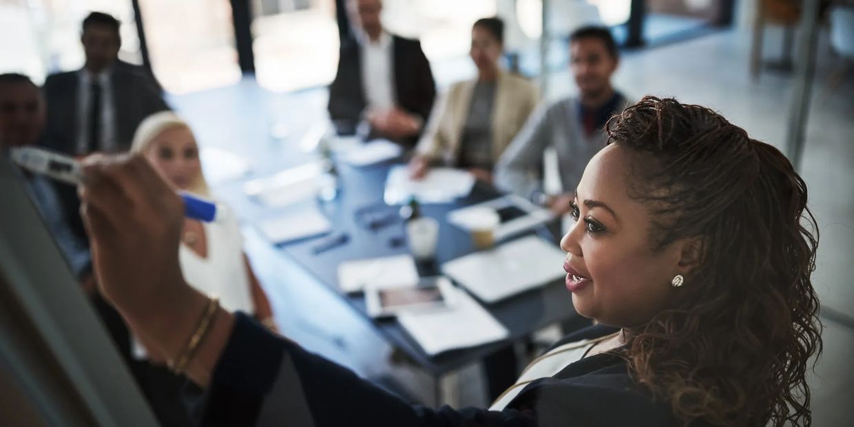 Woman leading a business meeting, writing on a whiteboard with colleagues attentively watching.