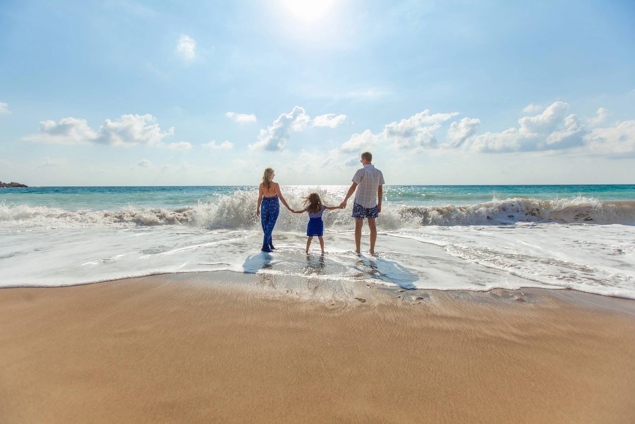 A family of three holding hands and facing the ocean on a sunny beach.