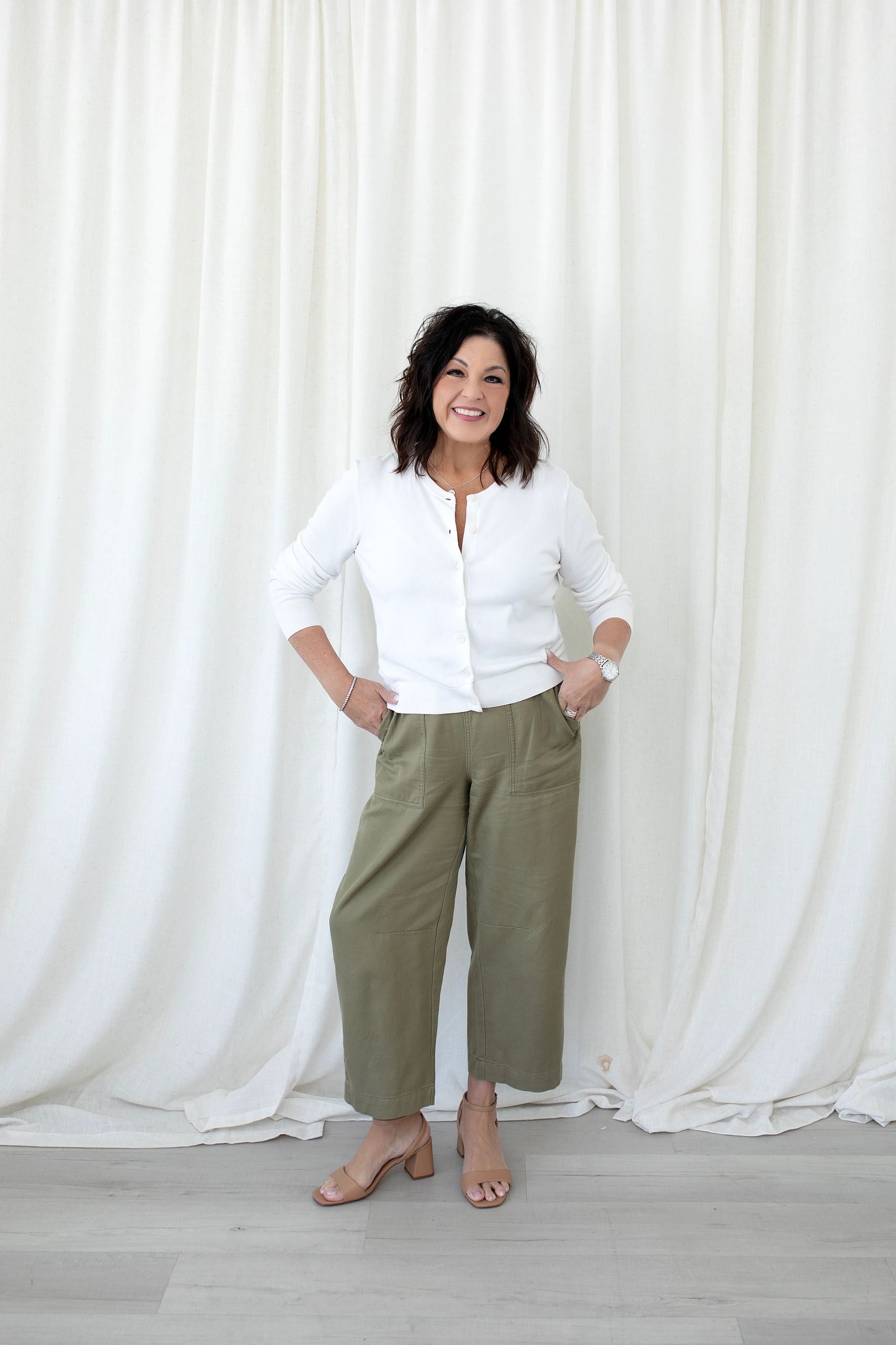 Smiling woman in white sweater and olive pants against a white curtain backdrop.