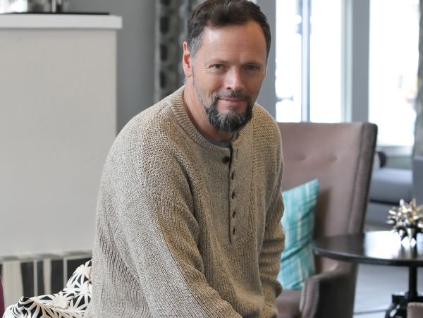 A man with a beard wearing a knitted sweater, sitting indoors and smiling.
