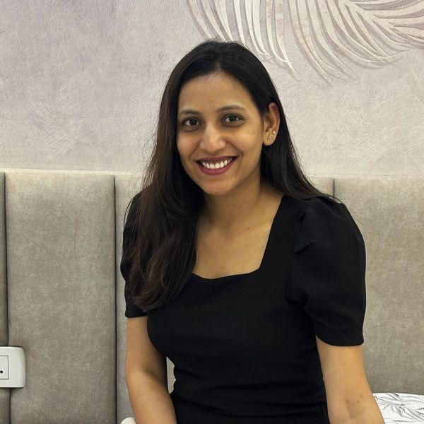 Woman sitting on a bed smiling, dressed in a black top and beige pants.
