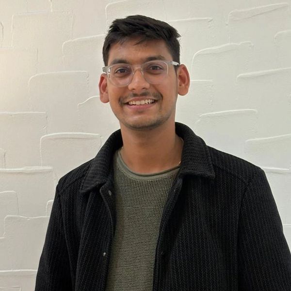 A young man smiling, wearing glasses, a black jacket, and a green sweater.