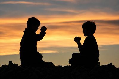 Two children praying together at sunset, silhouetted against a colorful sky.
