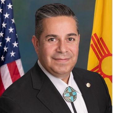 Portrait of a man in a suit with American and New Mexico flags in the background.