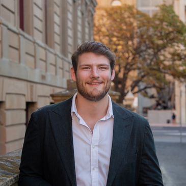 Smiling man in a blazer standing on a city street with autumn trees in the background.