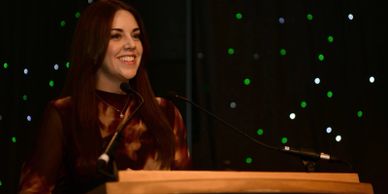 A woman smiles while speaking at a podium with a dark, starry background.
