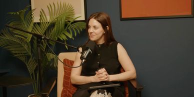 A woman in a black sleeveless top speaking into a microphone in a cozy studio.