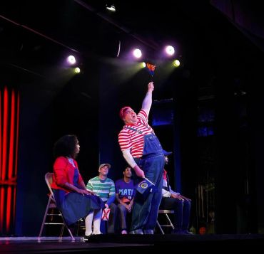 Performer in striped shirt entertaining on stage with audience seated.