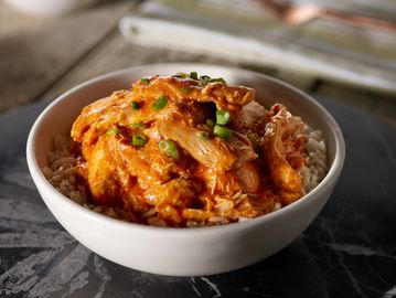 Big bowl of shredded chunks of chicken, buffalo sauce orange in color, red pepper flakes, over rice.