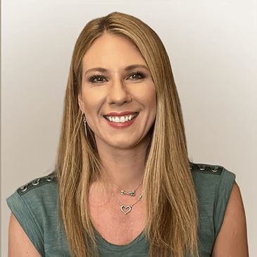 A smiling woman with long blonde hair wearing a green top and heart necklace.