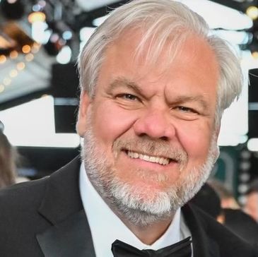 Smiling elderly man with white hair and beard in a tuxedo at an event.