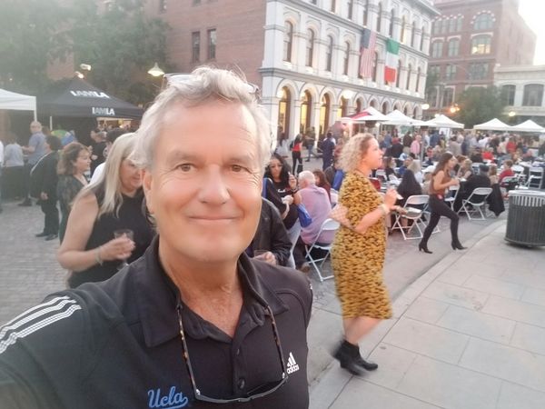 Man taking a selfie at an outdoor event with people seated and socializing.