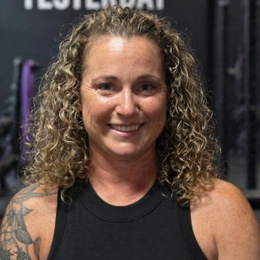 Smiling woman with curly hair and a tattoo wearing a black sleeveless top.