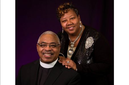 Smiling couple dressed formally, with the man wearing clerical attire and the woman adorned with elegant jewelry.
