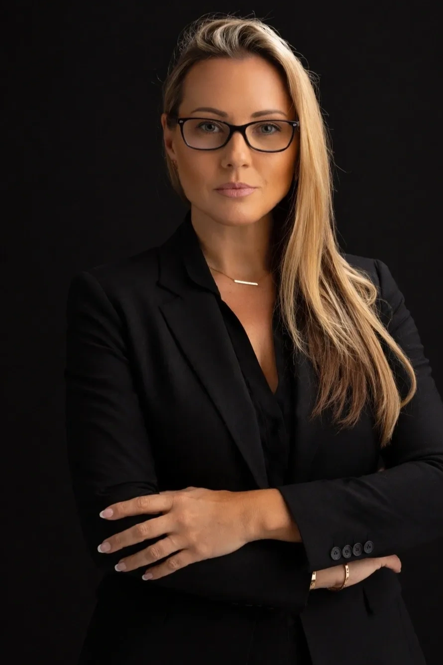 Confident woman in black suit with glasses poses against dark background.