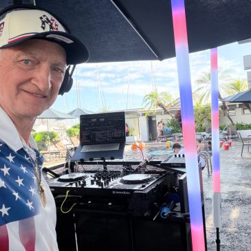DJ in American flag shirt with DJ equipment at outdoor event.