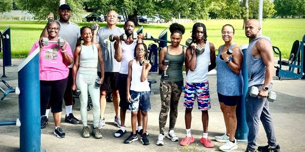 A diverse group holding dumbbells at an outdoor gym, smiling and posing for a photo.