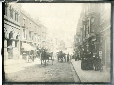 Black and white photo of a street scene with horse carriages and pedestrians.