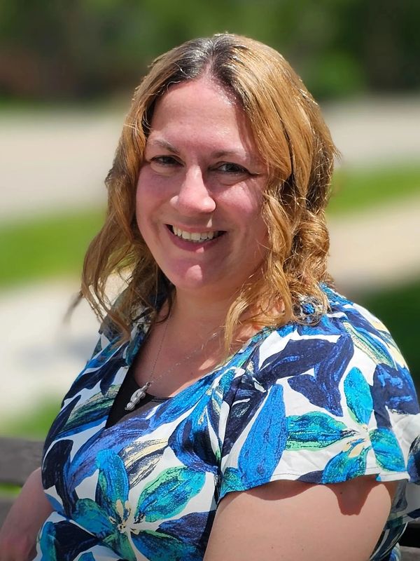 Smiling woman with curly hair wearing a blue floral top outdoors.
