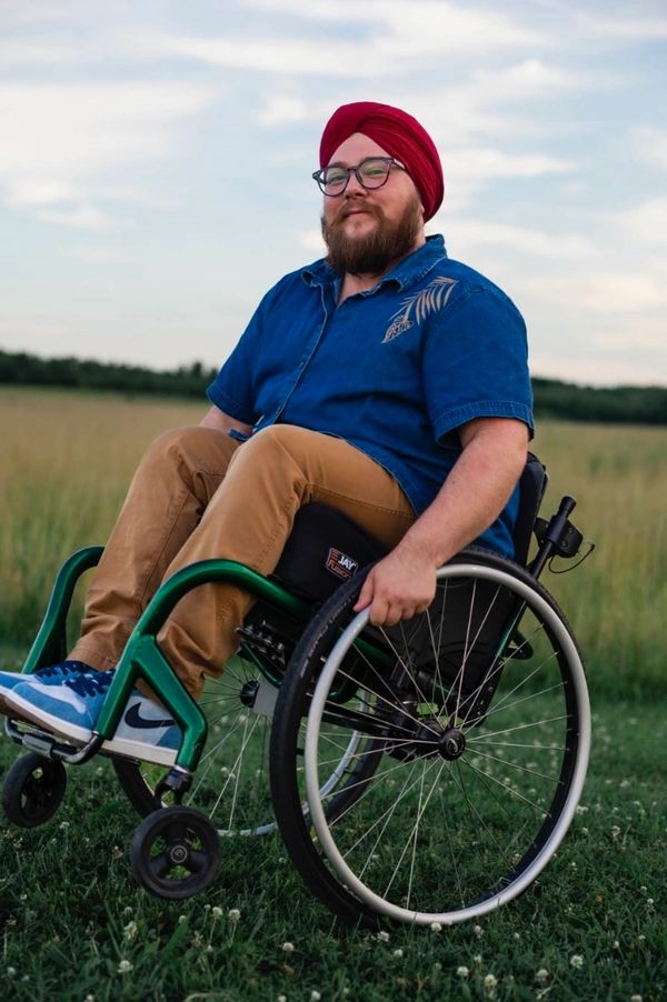 Jordyn sitting in his wheelchair wearing glasses, a red turban, blue shirt, khakis, and blue shoes