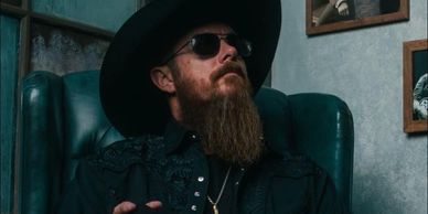 Man with a beard wearing sunglasses and a hat, sitting confidently.