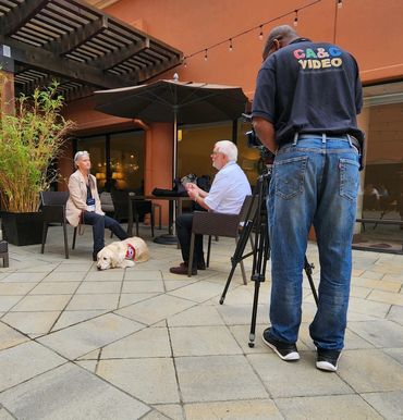 A woman in a business suit sits with a service dog at her feet. She is being interviewed on camera