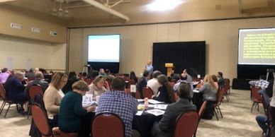 People attending a conference or workshop in a large room with presentations.