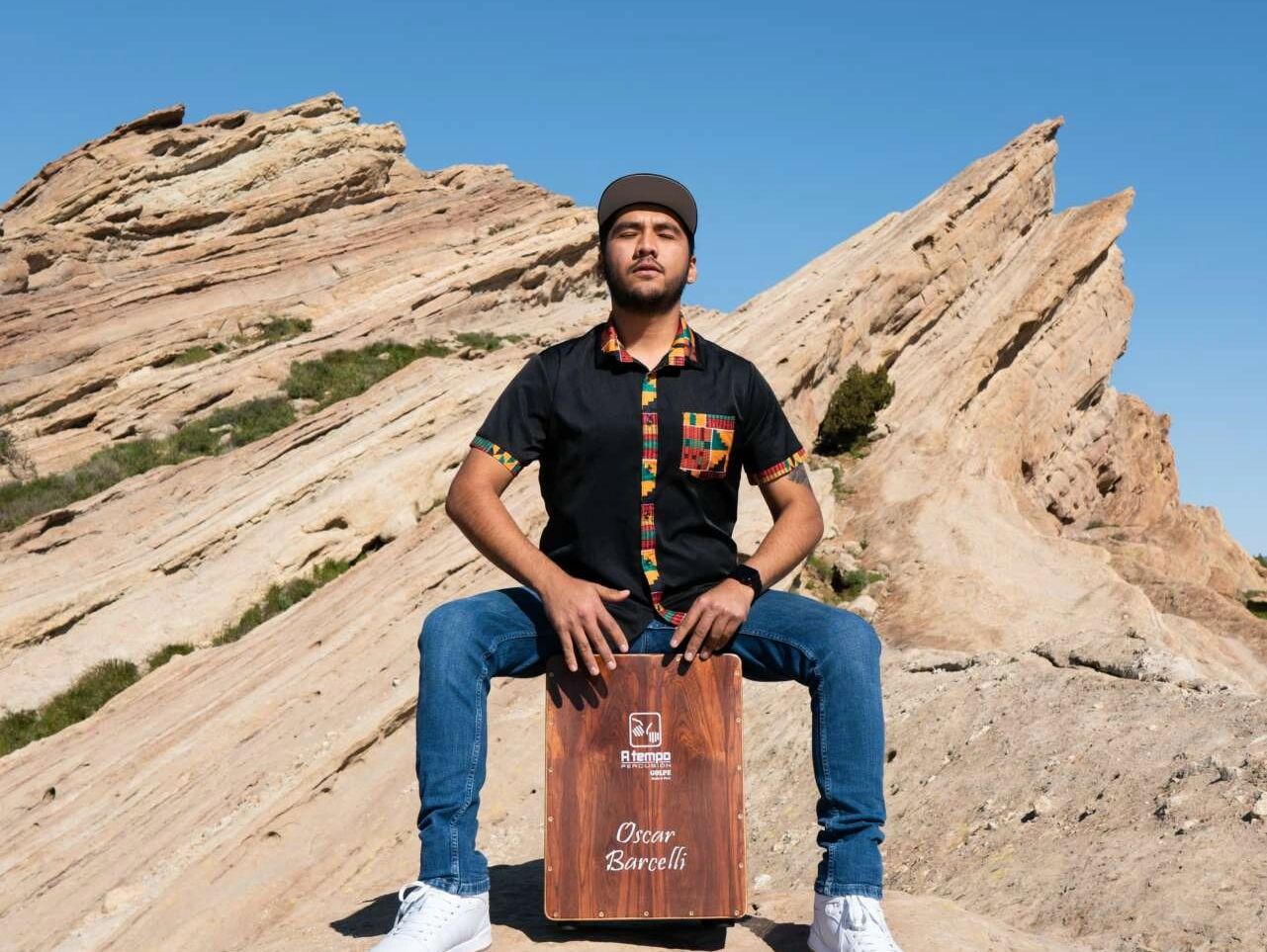 Man playing a wooden cajón drum outdoors against rocky formations.